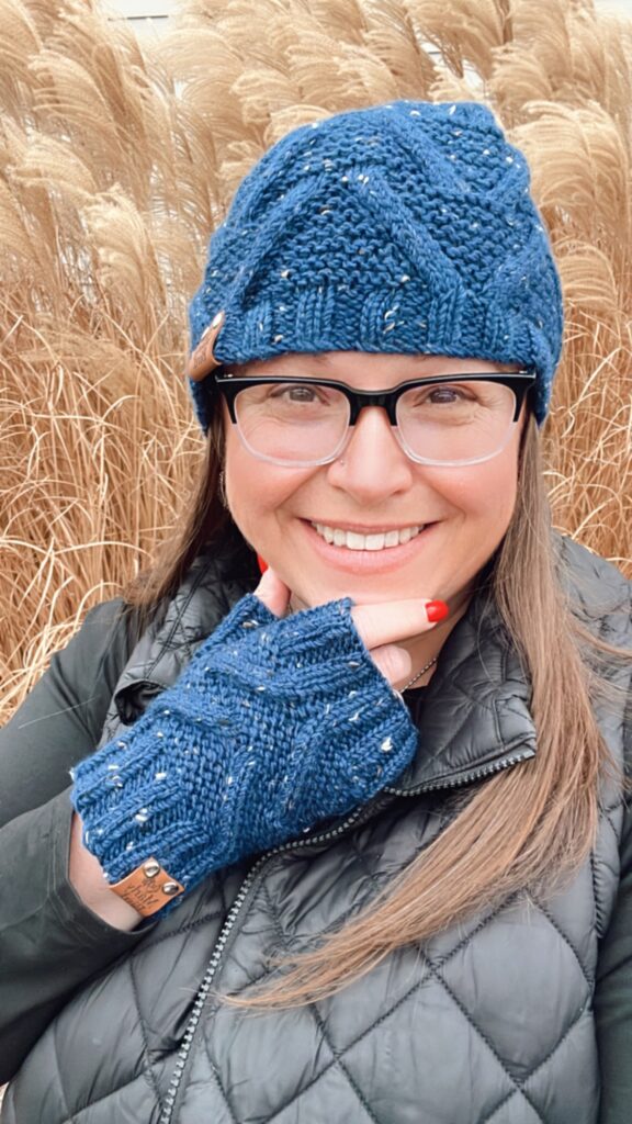 Marly Bird smiling, wearing the 'Take a Look' knit cabled hat and matching fingerless mittens in a rich blue tweed. The hat is topped with a fluffy blue pompom, and the mittens feature intricate cable patterns. She is posing against a backdrop of tall, golden reed grass.