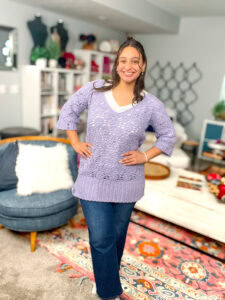 Woman models a lavender knit sweater with visible stitch texture in a bright room with crochet home decor and patterned rug.