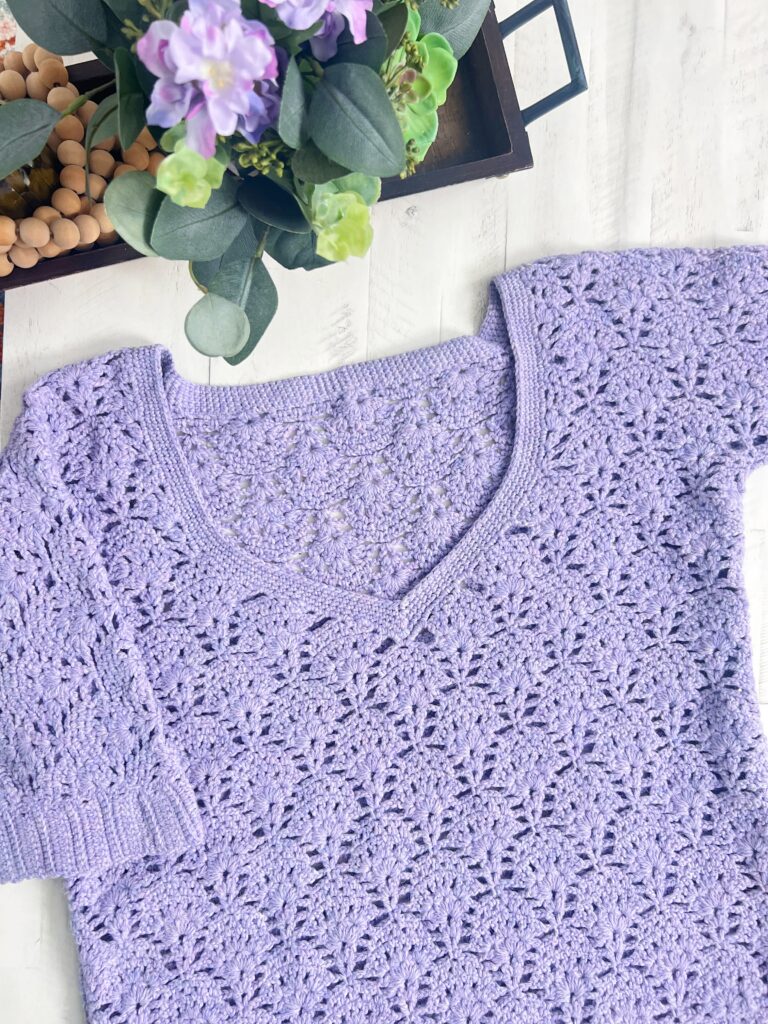 A light purple Alpaca Crochet Sweater Pattern short-sleeve top with a V-neck and intricate floral patterns is laid flat on a white surface next to a tray with a potted plant and wooden beads. -Marly Bird