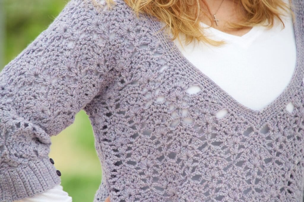 A close-up of a person wearing a white shirt under a lavender, floral-patterned Alpaca Crochet Sweater Pattern. The person’s face is not visible; only part of their arm and shoulder are shown. -Marly Bird