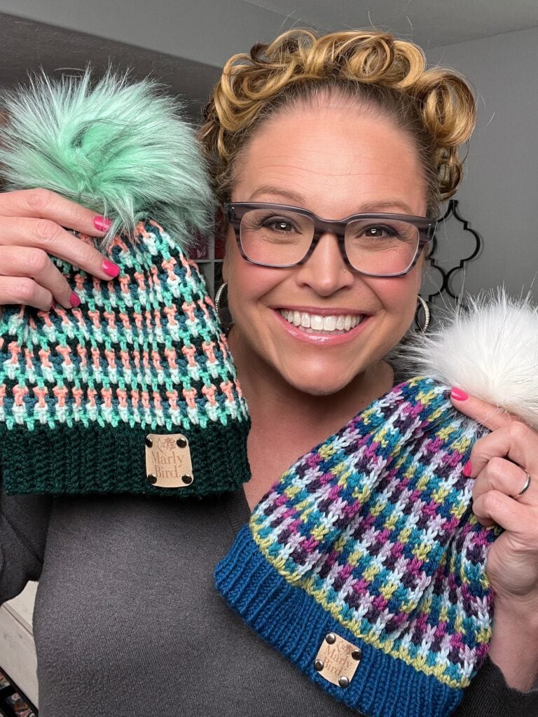 A smiling person with glasses and curly hair holds up two colorful stash-busting knit hats with pom-poms, one green and pastel, the other blue and multicolored, each showcasing a "Marly Bird" tag. -Marly Bird