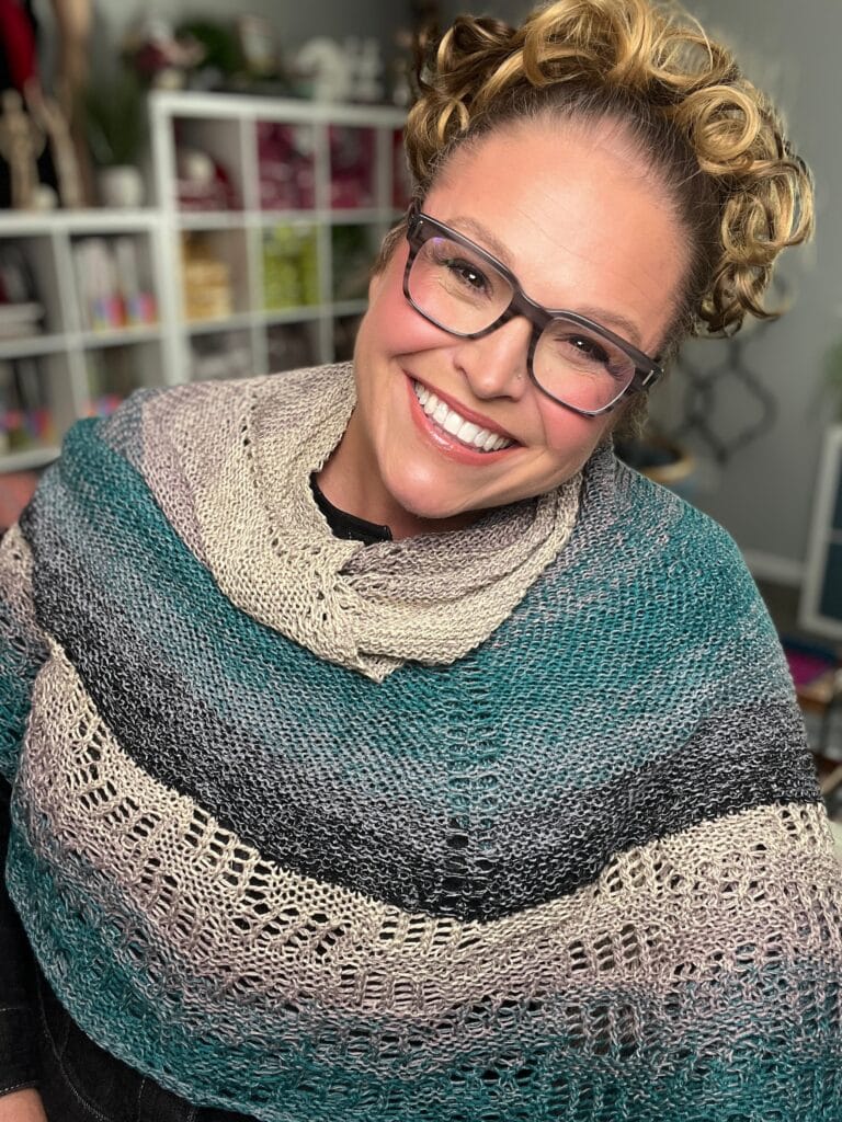 Teal, gray, and beige Outlander knit shawl with textured stitches, modeled indoors by a woman with curly hair and glasses.