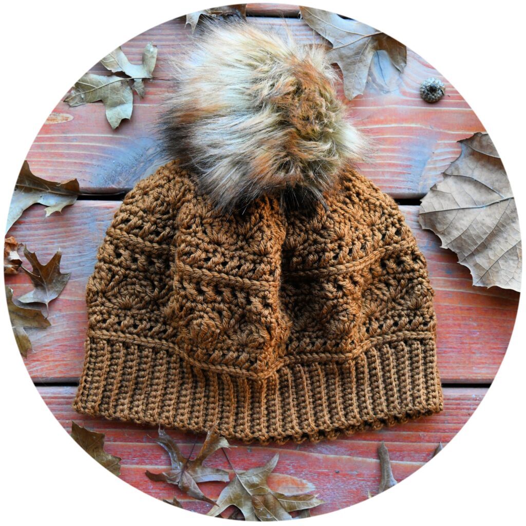 Knit-Look Crochet Hat with a large, fluffy pom-pom on top is displayed on a wooden surface surrounded by scattered dried leaves. The beanie features intricate stitching and ribbed edges. -Marly Bird