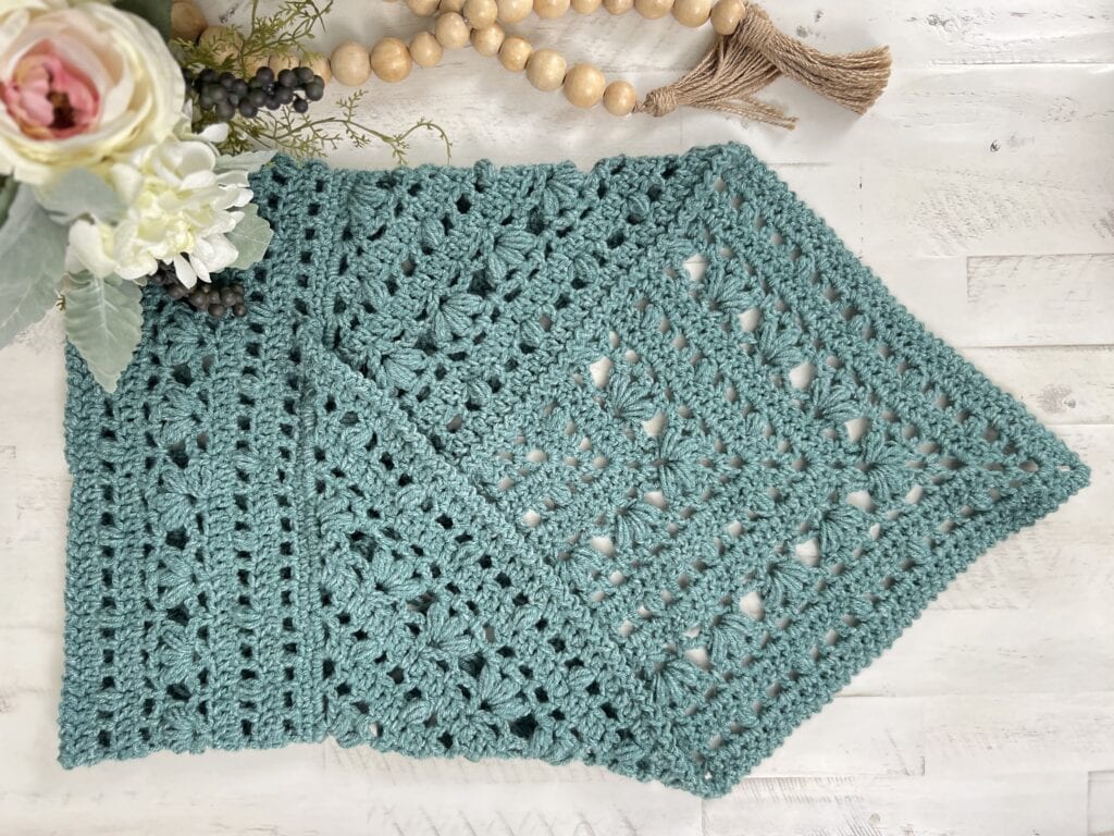 A teal crochet wrap with intricate patterns is gracefully displayed on a white wooden surface. Beside it, a floral arrangement and wooden beads enhance its charm, crafting an elegant and cozy Kembleford aesthetic reminiscent of a handmade cowl. -Marly Bird