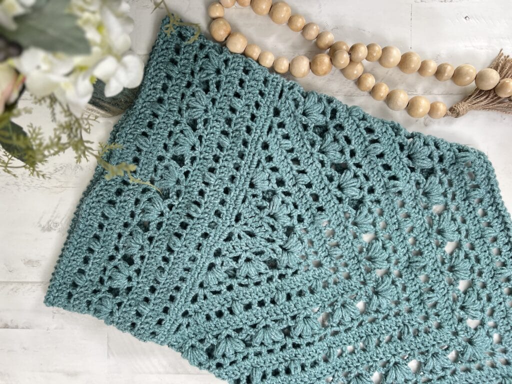 A close-up of a crocheted teal blanket, evoking the charm of Kembleford, features intricate patterns and textures. Beside it, a string of wooden beads and a hint of white flowers adorn the rustic white wooden surface. -Marly Bird