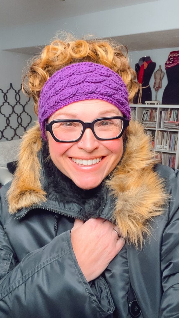 A smiling person with curly hair wears black glasses, a purple knit headband, and a winter coat with a faux fur collar. Bookshelves and mannequins are visible in the background. -Marly Bird