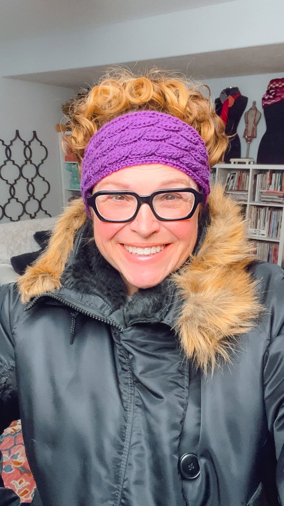 Smiling person with curly hair wears a purple knit headband, black glasses, and a black coat with a furry collar; they are indoors with bookshelves and mannequins visible in the background. -Marly Bird
