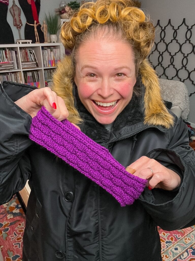 A smiling woman in a black coat with a fur-trimmed hood and curlers in her hair holds up a purple knitted headband. She stands indoors with shelves and books visible in the background. -Marly Bird