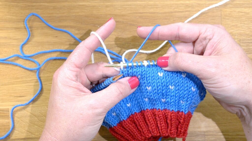 The image depicts a close-up of a person's hands performing the tucking in or catching  floats technique in stranded knitting. The hands are shown holding knitting needles and yarn, with the left needle displaying a work in progress that features two colors: blue and white. The person's right hand is using a knitting needle to manipulate the blue yarn, while the left hand is controlling the tension of the white yarn, which is being carried or 'floated' across the back of the work along the bottom. This technique is used to manage the long strands of yarn on the back side of colorwork knitting, ensuring that the floats are secured and the tension remains even. The image clearly shows the intricate process of managing yarns during colorwork knitting, a skill necessary for creating neat and even fabric in multi-color patterns. This is also a depiction of holding the color dominant yarn in the left hand and the non-dominant yarn  color in the right had as a knitter does stranded knitting - Marly Bird





