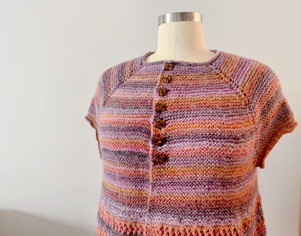 A hand-knitted short-sleeve cardigan displayed on a mannequin. The cardigan showcases a beautiful gradient of warm hues, ranging from soft lavender to deeper shades of purple and peach. It features a delicate ribbed neckline, a vertical button band with decorative buttons down the front, and a subtle textured edging. The piece is crafted with a lightweight yarn that creates a soft drape, suitable for a comfortable, yet stylish look.A hand-knitted short-sleeve cardigan displayed on a mannequin. The cardigan showcases a beautiful gradient of warm hues, ranging from soft lavender to deeper shades of purple and peach. It features a delicate ribbed neckline, a vertical button band with decorative buttons down the front, and a subtle textured edging. The piece is crafted with a lightweight yarn that creates a soft drape, suitable for a comfortable, yet stylish look.