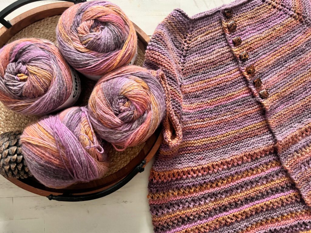 Image showcases the 'Bewitch' color variant of Caron Macchiato Cakes yarn, arranged in a rustic woven basket beside the completed 2XL size Shoop Shoop Shoop Knit Cardigan by Marly Bird, part of the Turkey Trot Make-Along 2023. The yarn features a mesmerizing palette of purples, pinks, and oranges, reminiscent of a sunset. The cardigan laid flat reveals a gradient stripe pattern with delicate button details along the edge, highlighting the yarn's transition of colors and the cozy, handcrafted quality of the garment.