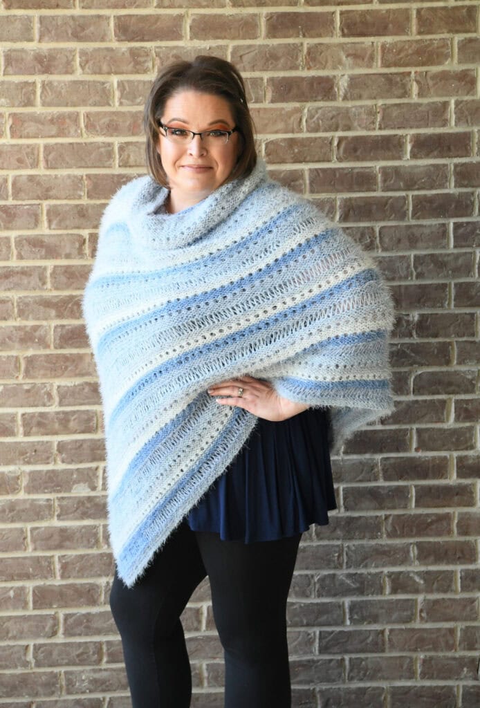 A woman with glasses stands in front of a brick wall, wearing a blue and white striped shawl—shown here in both knit and crochet versions of the same pattern—over a dark tunic and black leggings, hand on hip and a slight smile. -Marly Bird