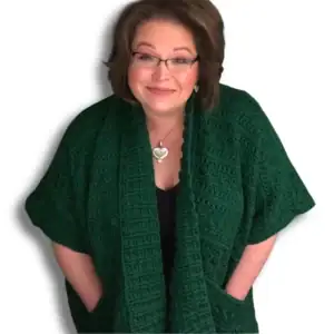 A woman with brown hair and glasses smiles warmly, wearing a dark green, textured crochet cardigan made from free knitting and crochet patterns, with front pockets and a heart-shaped necklace, standing against a white background. -Marly Bird