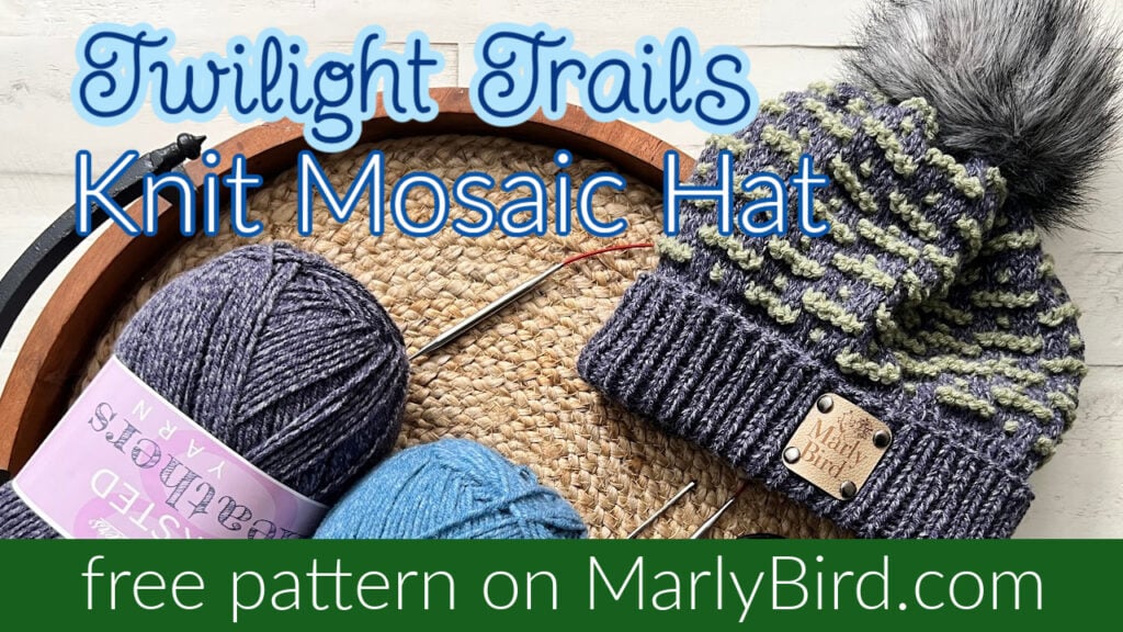 A knitted mosaic hat with a pom-pom sits on a tray surrounded by balls of yarn and knitting needles. The text reads "Twilight Trails Knit Mosaic Hat Pattern" and "free pattern on MarlyBird.com," all set against a light background. -Marly Bird