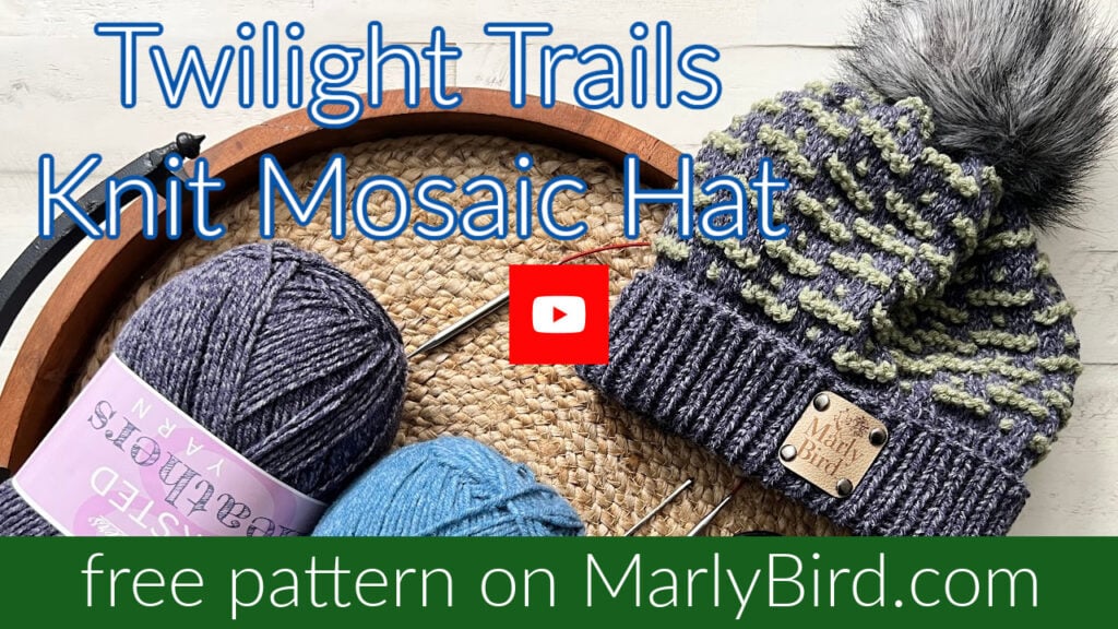 A knitted mosaic hat with a stylish textured design and a pom-pom sits on a circular wooden tray. Beside it are two yarn balls in purple and blue. The text reads, "Twilight Trails Knit Mosaic Hat," with a YouTube logo, and "free pattern on MarlyBird.com" at the bottom. -Marly Bird