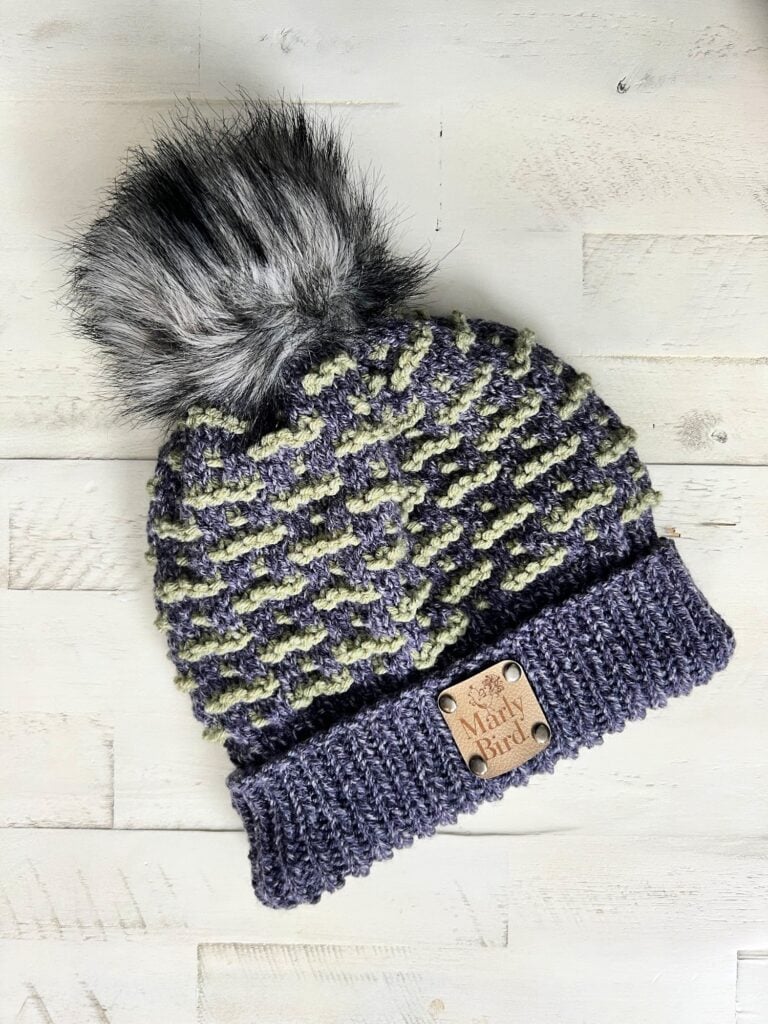 A knitted beanie, perfect for KNIT GAME DAY, features a grey and green textured pattern with a fluffy pom-pom on top. It has a ribbed brim adorned with a leather patch that reads "Marly Bird." The backdrop is a light wooden surface. -Marly Bird