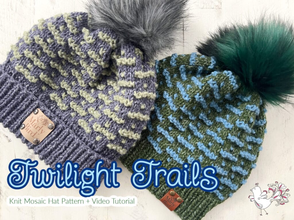 Two knit hats with vibrant color patterns and faux fur pom-poms are displayed. One is gray and green, the other blue and green. Each features a logo tag. Text reads "Twilight Trails" alongside a small bird graphic, highlighting the "Knit Mosaic Hat Pattern + Video Tutorial. -Marly Bird