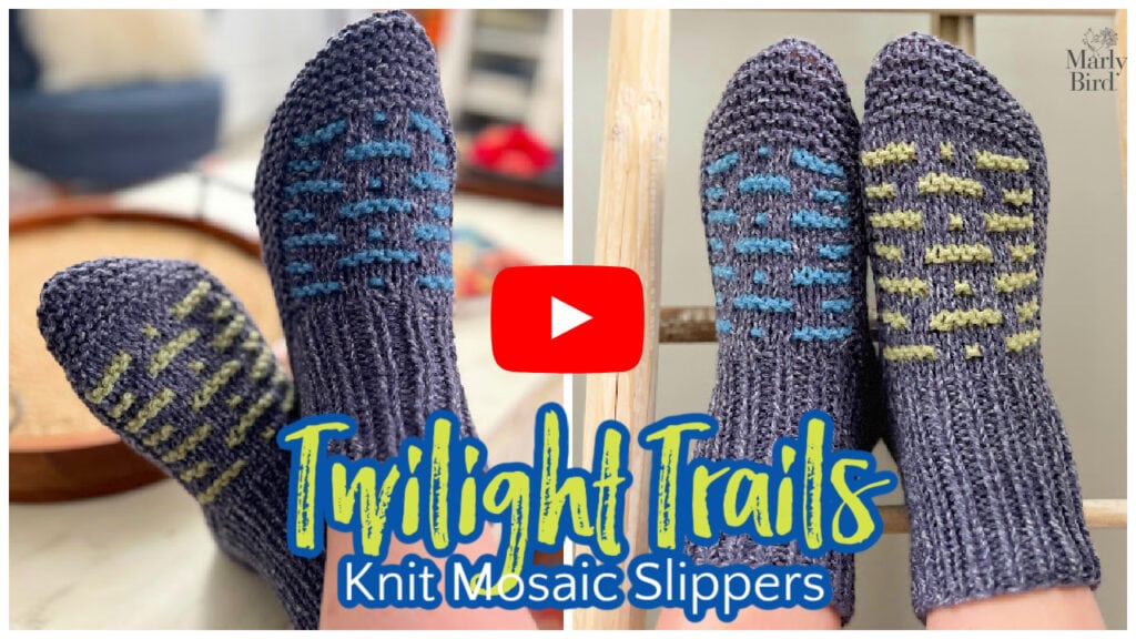 Close-up of two pairs of knit mosaic slippers in gray shades with blue and yellow patterns. The text "Twilight Trails Knit Mosaic Slippers" overlays the image, and a red play button sits in the center, inviting viewers to watch the video about these cozy creations. -Marly Bird