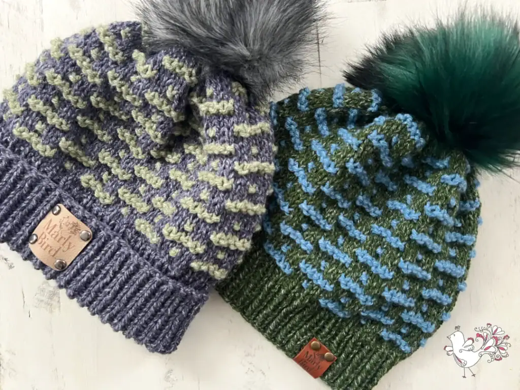 Two knitted beanies with faux fur pom-poms—one gray and light green, the other green and blue. Both feature a "Misty Yard" leather tag and rest on light wood. A bird logo appears in the corner, evoking cozy inspiration for free knitting and crochet patterns. -Marly Bird