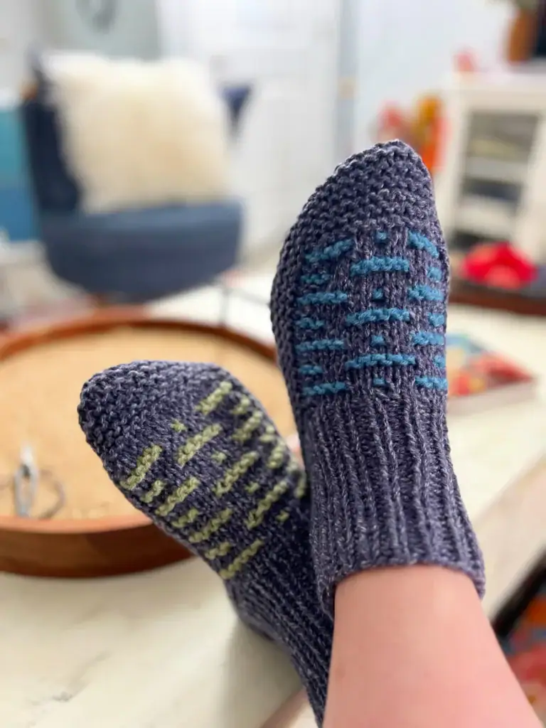 Close-up of hand-knit gray socks with blue and yellow patterning, showing textured stitches as worn on relaxed feet.