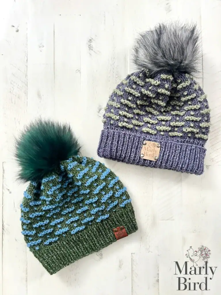 Two knitted beanies with pom-poms sit on a white wood background. Featuring woven labels and the "Marly Bird" logo, these hats showcase colors and style inspired by free knitting and crochet patterns. -Marly Bird