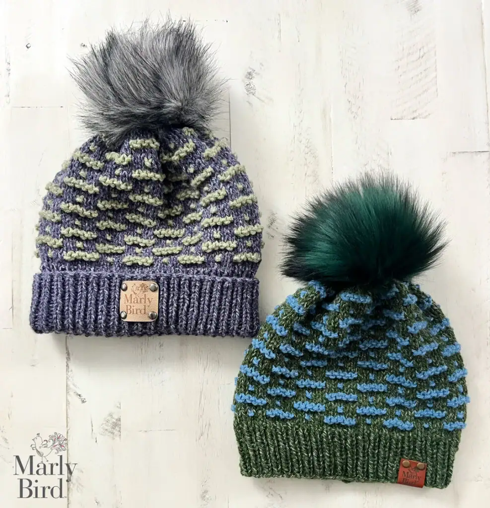 Two knit beanies with fluffy pom-poms—one gray and lavender, the other green and blue—both with branded leather tags, rest on a white wooden surface. Find inspiration in these colors for your own free knitting and crochet patterns. -Marly Bird