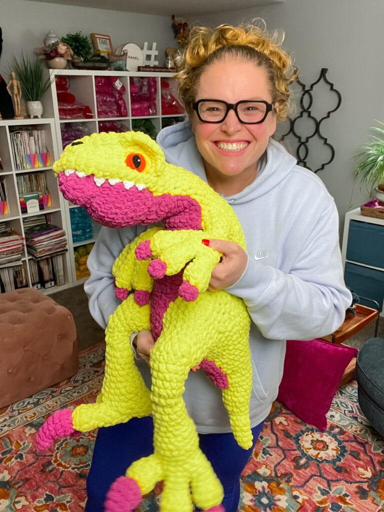 A smiling person with curly hair and glasses holds a bright yellow and pink dinosaur toy in a cozy, colorful room filled with shelves, books, and decorative items—perfect inspiration for those seeking free knitting and crochet patterns. -Marly Bird