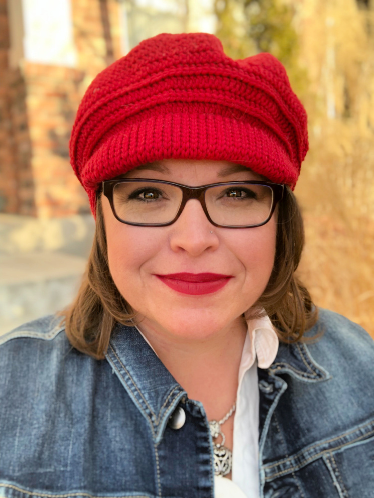 A woman is smiling at the camera, wearing a bright red ribbed crochet hat that fits snugly on her head. She has short brown hair, glasses, and red lipstick that matches her hat. She is also wearing a white shirt under a blue denim jacket, and a silver necklace with a heart-shaped pendant. The background is outdoors with blurred autumnal trees and a brick structure, giving the image a warm, seasonal feel. The woman appears to be in a happy, cheerful mood. - marly bird valentina crochet hat with bill
