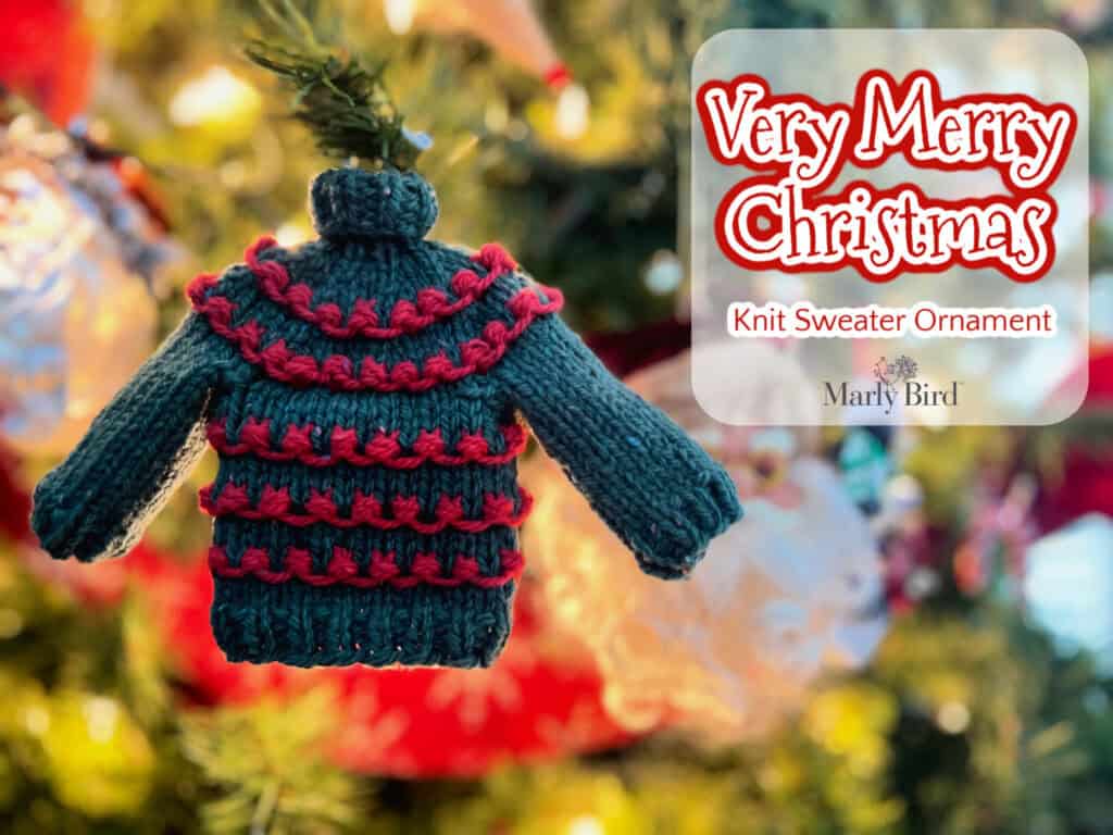 A small green knit sweater ornament with red accents hangs on a Christmas tree, offering a festive flair. Text reads, "Very Merry Christmas, Knit Sweater Ornament, Marly Bird." The background sparkles with twinkling lights and blurred ornaments. Discover the free pattern for your own holiday creation. -Marly Bird