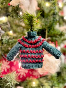 A charming miniature knitted sweater ornament with green and red stripes hangs on a Christmas tree, illuminated by twinkling lights and accompanied by a white snowflake ornament in the background. Discover the free pattern for your own Christmas knit sweater ornament to add extra delight. -Marly Bird