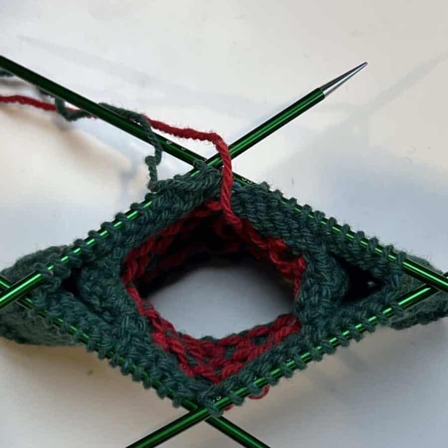 Close-up of green knitting in progress on double-pointed needles, forming a square shape, reminiscent of a festive knit sweater. Red yarn accentuates the central opening, with ends hanging loosely. The arrangement forms a geometric pattern against a plain white background, hinting at a Christmas theme. -Marly Bird