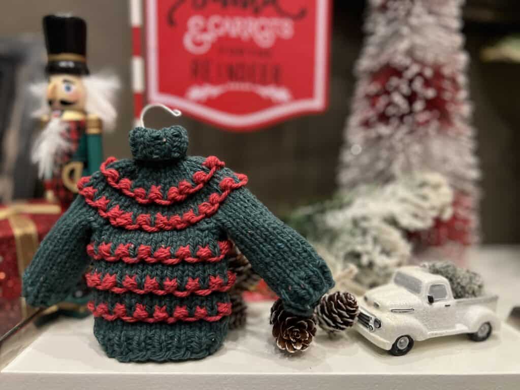 A miniature Christmas sweater with red and green stripes sits next to a small white toy truck and pinecones. In the background, a decorative nutcracker stands beautifully against red holiday-themed signage with blurred text, capturing the festive spirit. -Marly Bird