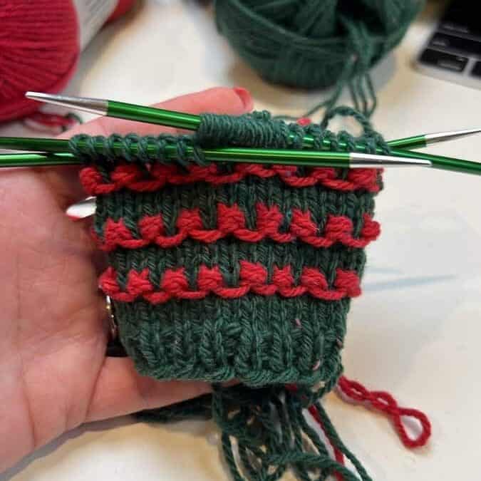 A hand holds green and red yarn knitting in progress, with knitting needles inserted into the fabric, crafting what seems like a festive Christmas knit sweater. Balls of matching yarn rest in the background on a white surface, ready to bring this free pattern to life. -Marly Bird