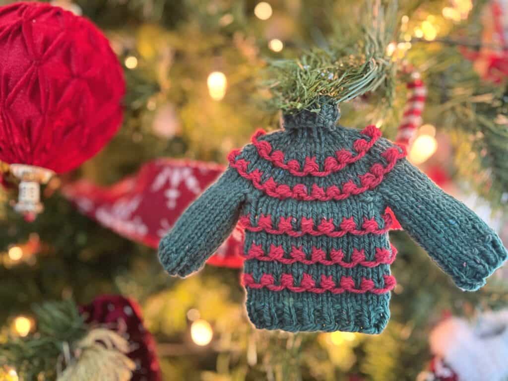 A green knitted sweater ornament with red detailing hangs on a Christmas tree, surrounded by twinkling lights and a large red ornament, capturing the cozy charm of the holiday season. -Marly Bird