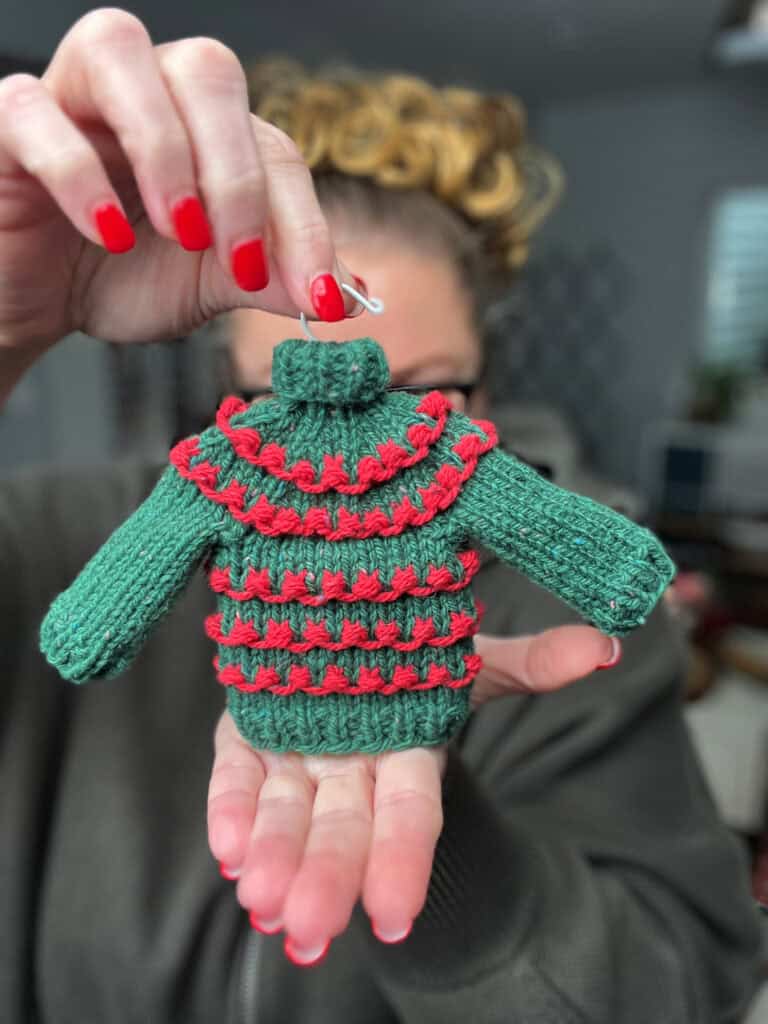 A person with red-painted nails holds a small green knitted sweater with red stripes, featuring a miniature hanger—a charming Christmas knit sweater ornament. The blurred background highlights this festive piece, perfect for a Very Merry Christmas atmosphere. -Marly Bird