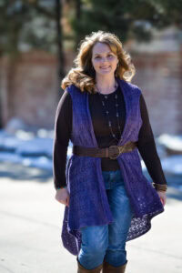 A woman models a long, purple knit lace duster, showing its openwork stitch detail and soft drape in natural sunlight.