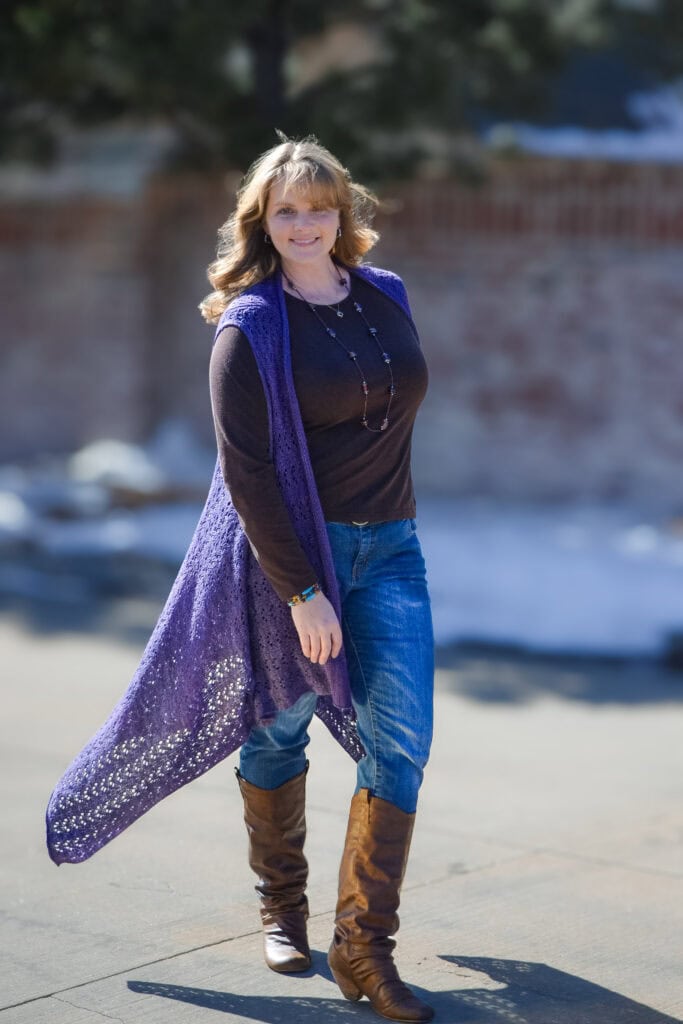 A woman models a hand-knit purple duster with an open front, textured stitch pattern, and relaxed drape over casual winter clothes.