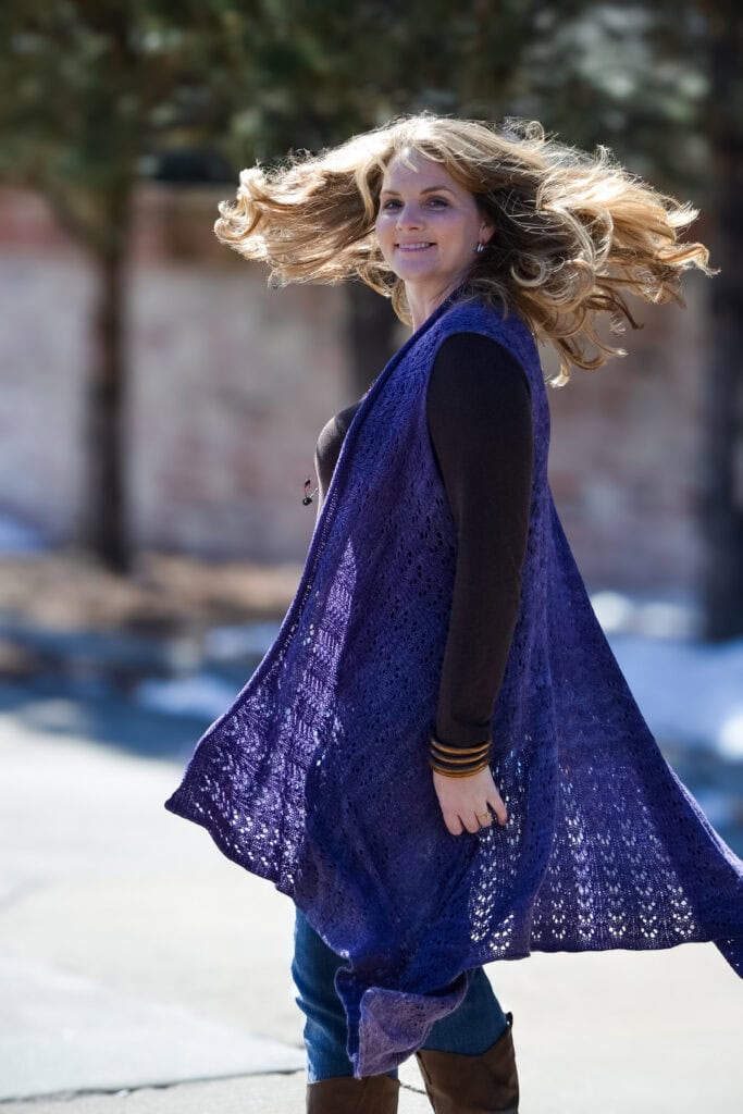 A woman models a long, open front knit duster with visible textured stitches over casual clothes, twirling outdoors.