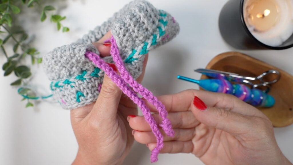 A person with red nail polish holds a gray crocheted item with teal accents and a pink braided cord. Nearby are a candle, plant, scissors, crochet hook, and a wooden tray. -Marly Bird