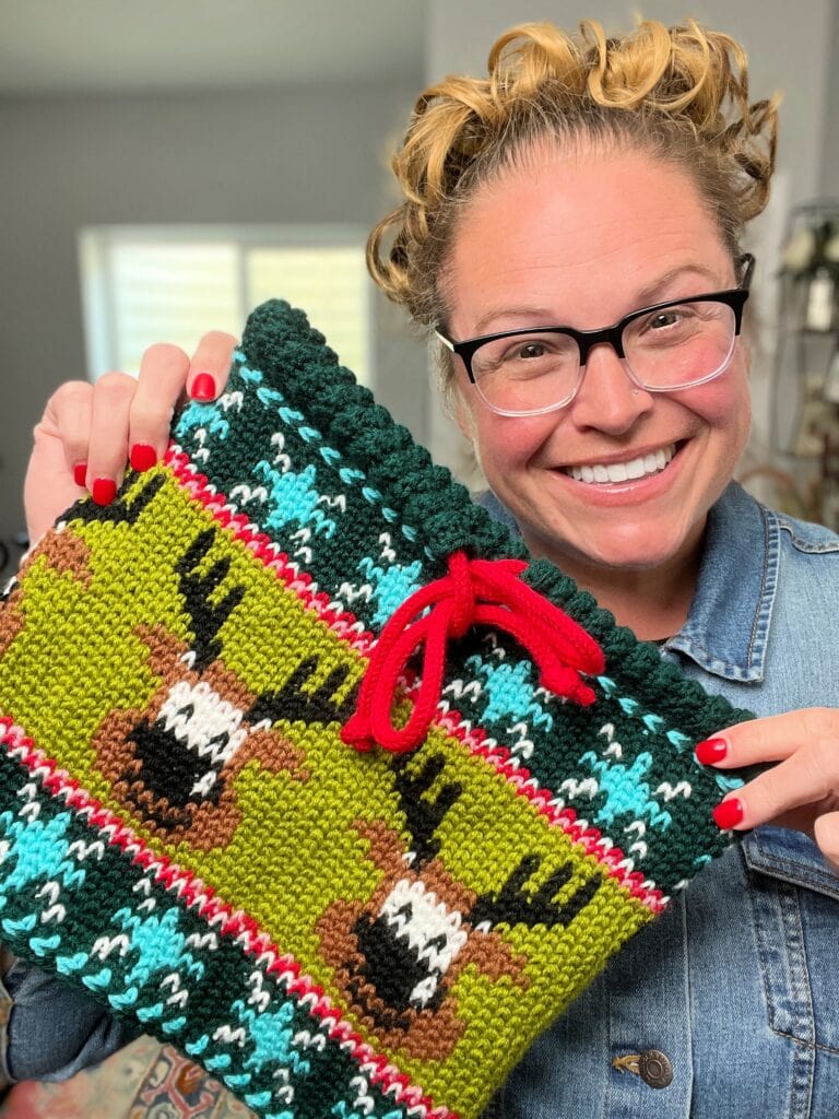 A smiling person with glasses and curly hair holds up a green, red, and blue knitted bag featuring reindeer and a red drawstring—showcasing festive Vixen’s Vibe. They have red-painted nails and are wearing a denim shirt. -Marly Bird