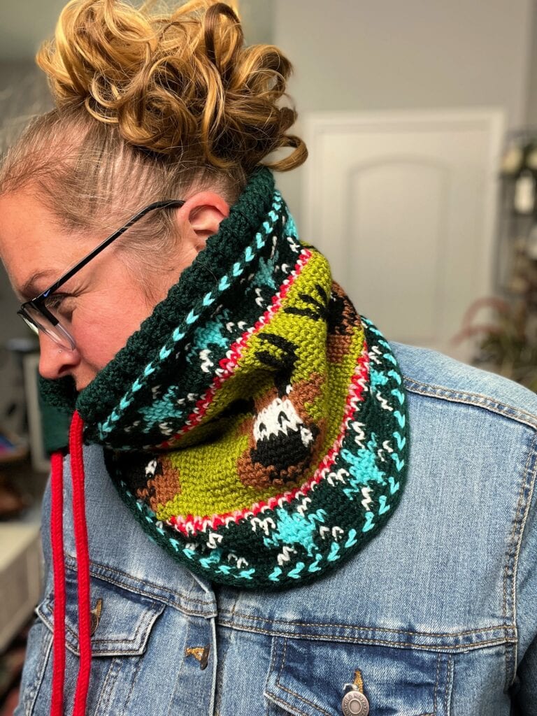 A person with curly hair and glasses wears a colorful Crochet Cowl called Vixen's Vibe, featuring a red drawstring and patterns of brown, green, turquoise, and white, over a denim jacket. The background is softly blurred. -Marly Bird