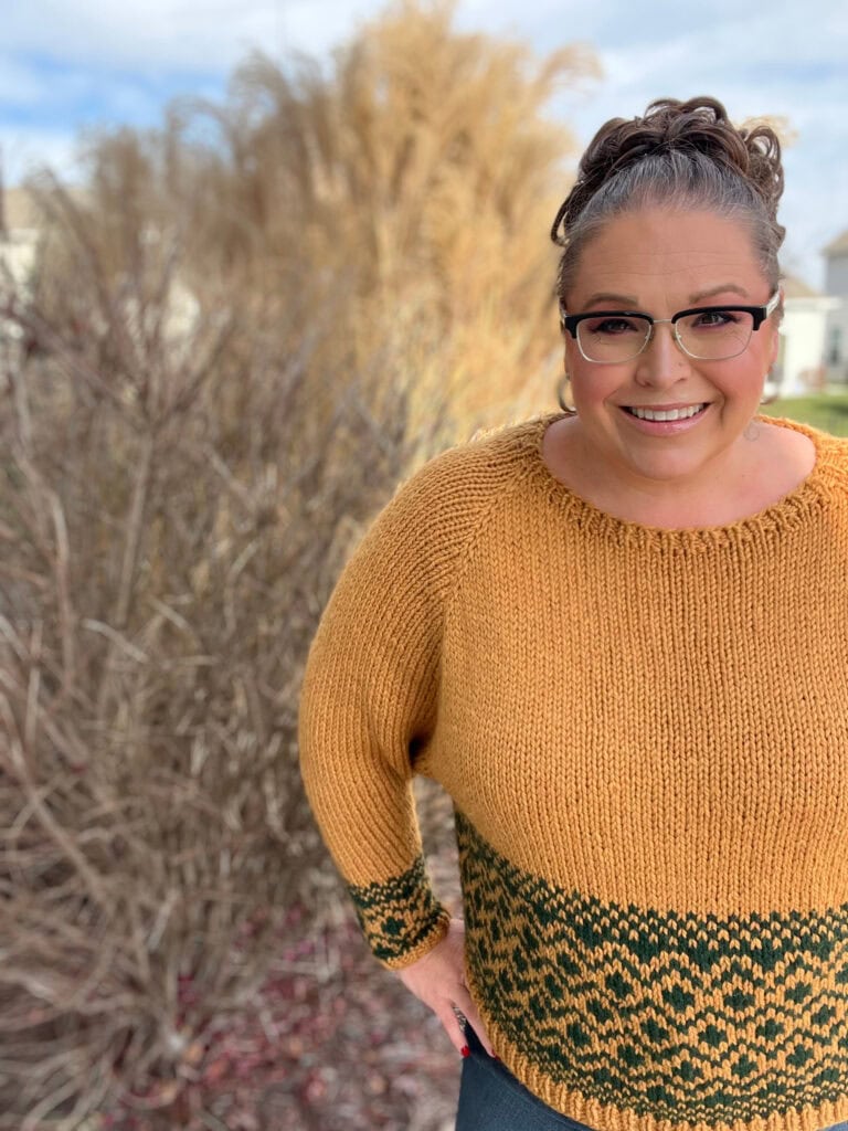 Woman modeling a hand-knit mustard sweater with green patterned band; visible texture and colorwork. Outdoor background.
