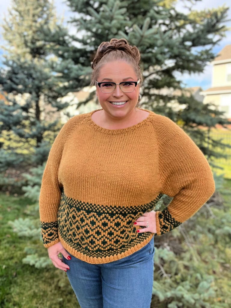 Woman outdoors models a hand-knit mustard sweater with dark green waist band, highlighting texture and colorwork detail.