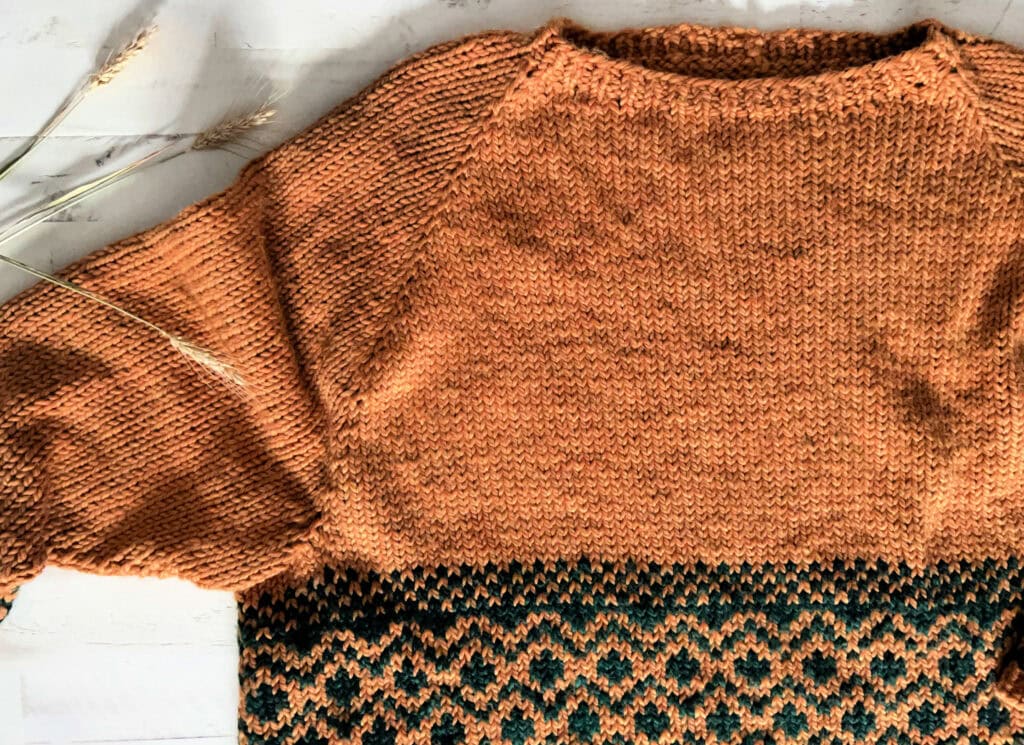 Close-up of an orange knit raglan sweater with a green and orange patterned hem. Dried grasses lie beside it on a pale surface.