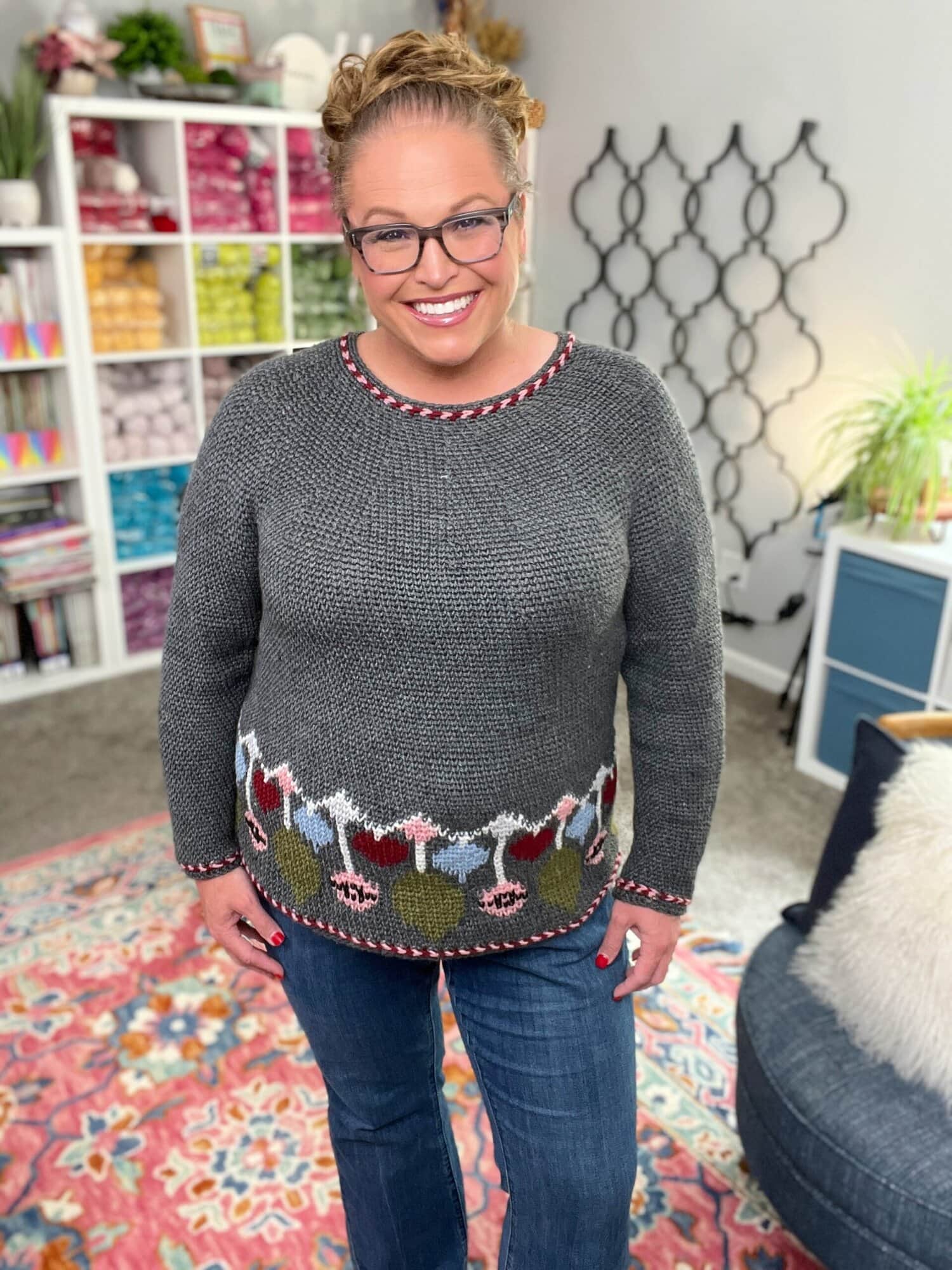 Gray knit sweater with Split SC Colorwork at the hem, shown worn in a yarn-filled, cozy room with decorative accents.