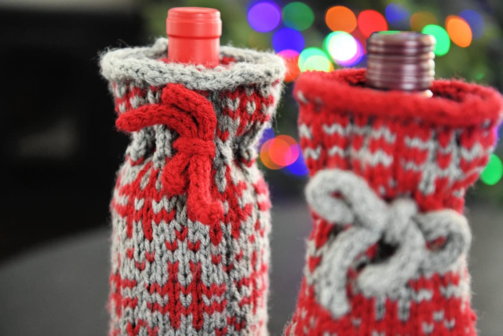 Two wine bottles wrapped in festive red and gray knitted wine cozies with bows. The background features colorful, blurred holiday lights, infusing the scene with a warm and cheerful atmosphere perfect for spreading holiday cheer. -Marly Bird