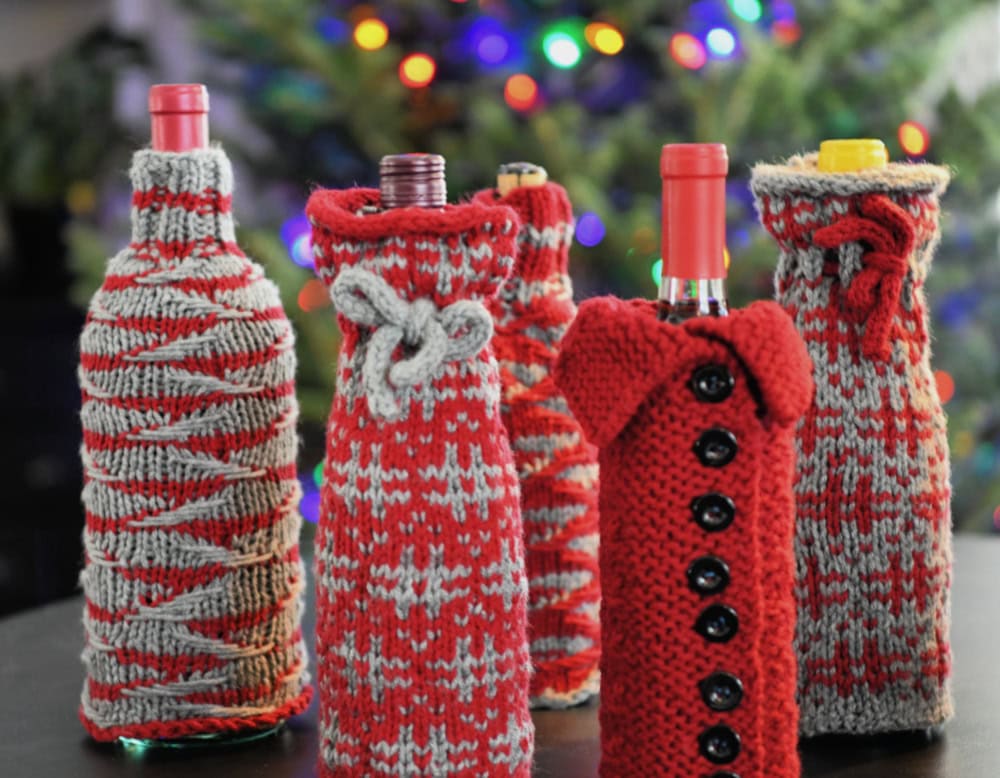 Festively decorated wine bottles wrapped in charming red and gray knit cozies with buttons, bows, and patterns. This delightful gift set evokes holiday cheer, as a blurred Christmas tree glittering with colorful lights creates a cozy atmosphere. -Marly Bird