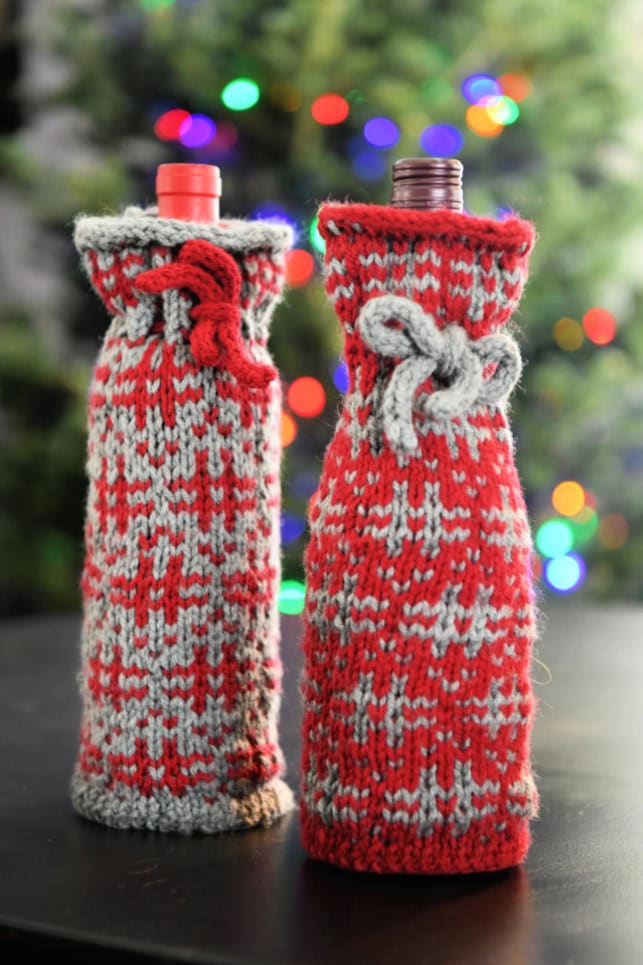 Two bottles adorned in red and gray knitted wine cozies with bow ties exude holiday cheer. Set on a black surface, they bask in the glow of blurred multicolored Christmas lights, making them the perfect gift kit for the festive season. -Marly Bird