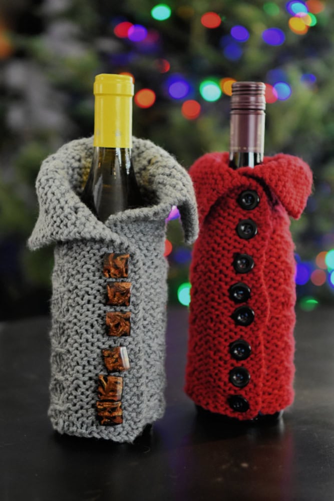 Two wine bottles, part of a Wine Cozy Triplets set, are dressed in knitted covers styled as winter coats—one in gray with toggle buttons and the other in red with black buttons. A blurred, colorful Christmas tree adds a touch of holiday cheer to the background. -Marly Bird