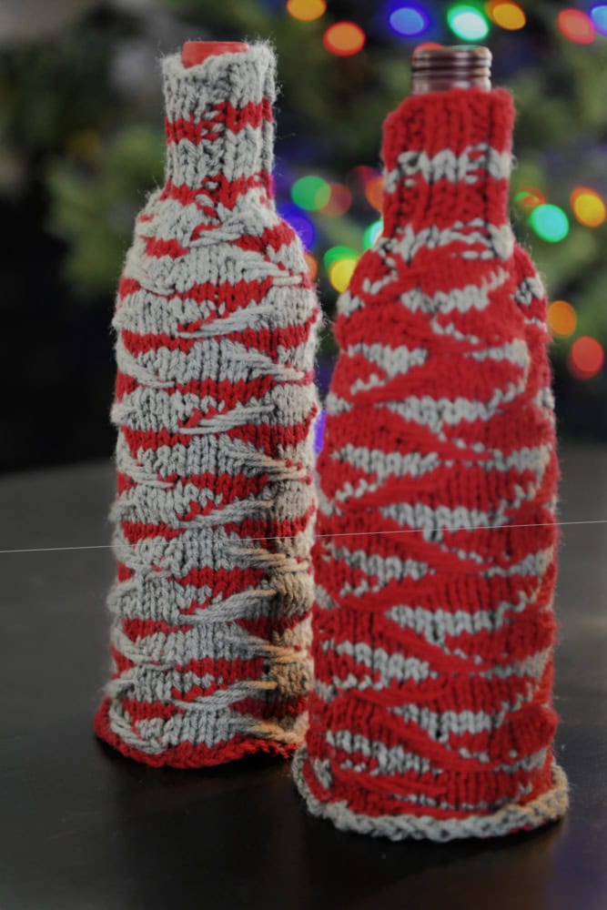 Two bottles wrapped in red and gray knitted wine cozies stand on a table. A colorful, blurred background with holiday lights creates a festive atmosphere, making it the perfect holiday cheer gift kit for any gathering. -Marly Bird